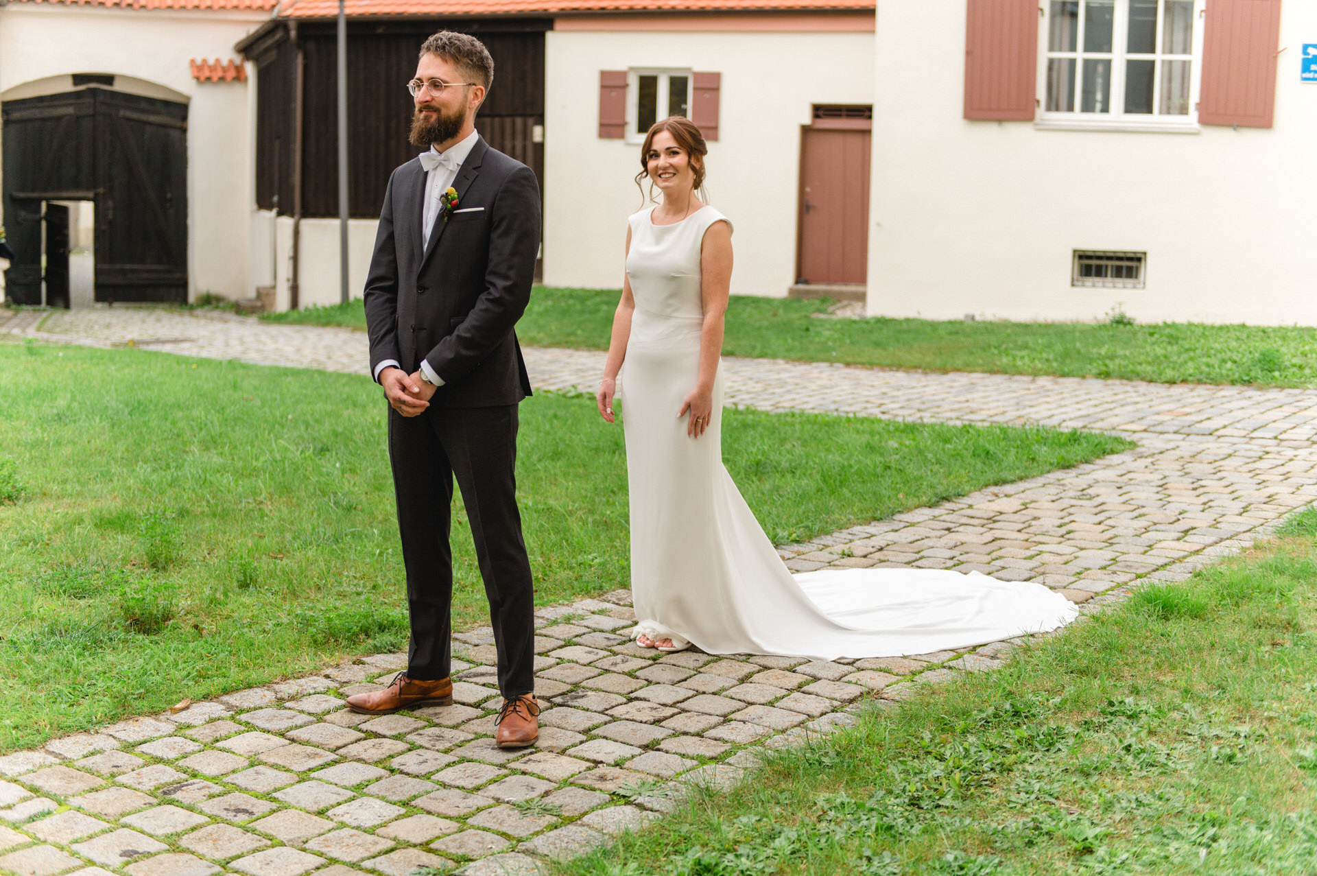 Eine Braut im weißen Kleid steht lächelnd in die Kamera, während der Bräutigam im Anzug vor der Kulisse eines historischen Gebäudes und eines Innenhofs wegschaut.