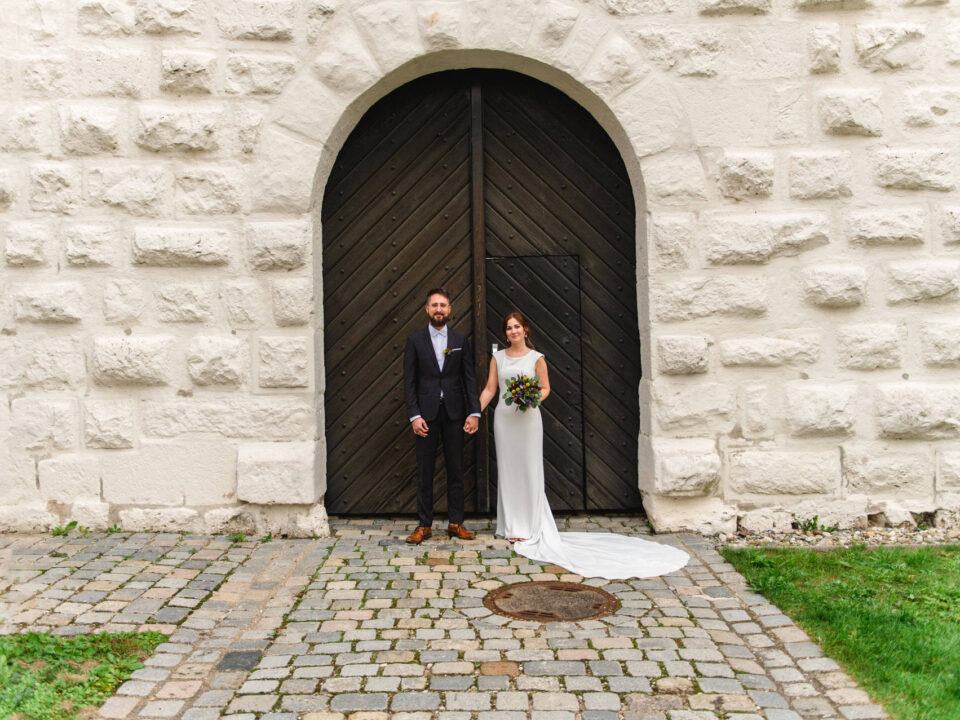 Eine Braut und ein Bräutigam stehen zusammen vor einer großen, gewölbten Holztür in einer weißen Steinmauer, halten sich an den Händen und lächeln.