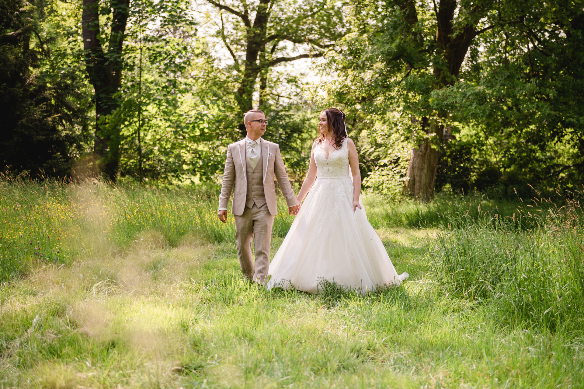 Ein Paar im Hochzeitskleid hält Händchen, während es durch eine von Bäumen umgebene Wiese geht.