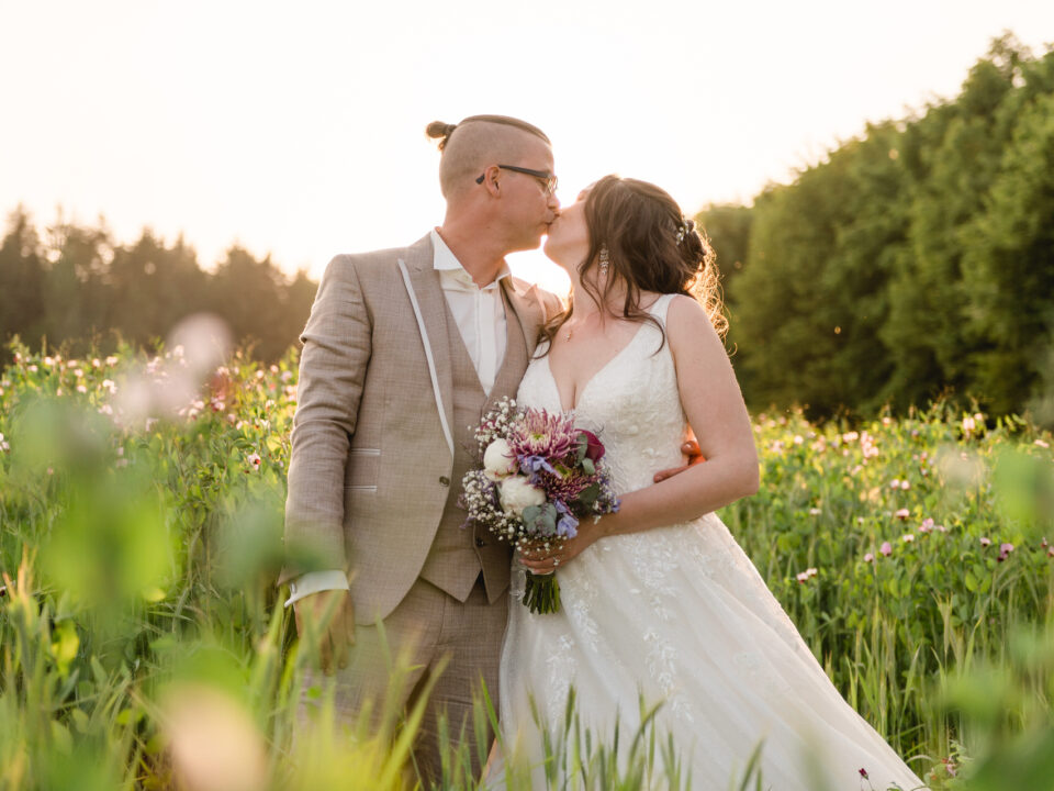 Ein Paar im Hochzeitskleid steht bei Sonnenuntergang auf einem Feld und küsst sich. Die Braut hält einen Blumenstrauß.