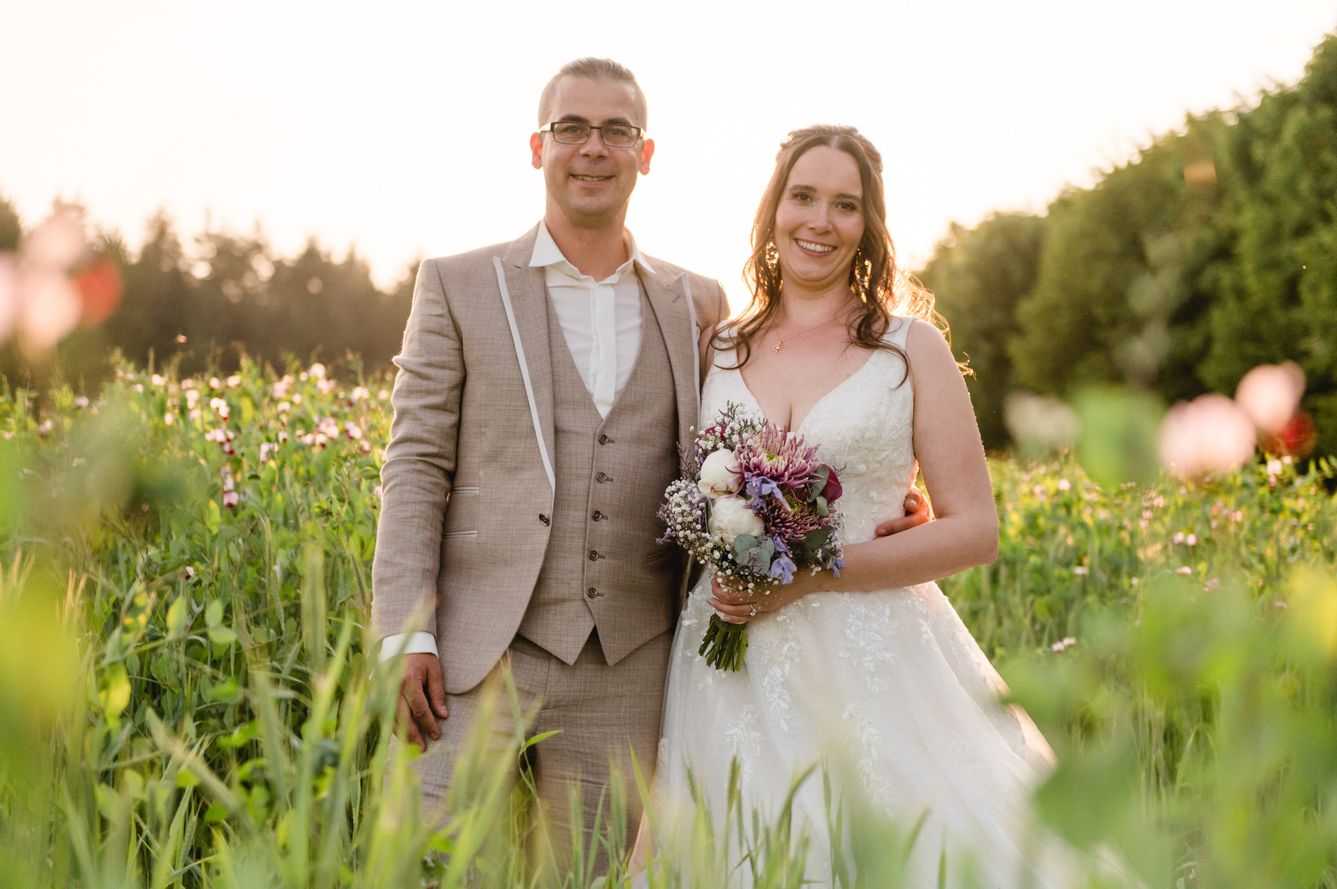 Eine Braut und ein Bräutigam stehen lächelnd bei Sonnenuntergang auf einer Wiese. Die Braut hält einen Blumenstrauß in der Hand.