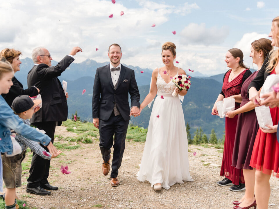 Braut und Bräutigam gehen Hand in Hand im Freien, während die Gäste Blütenblätter nach ihnen werfen. Im Hintergrund sind Berge zu sehen und die Gruppe lächelt.