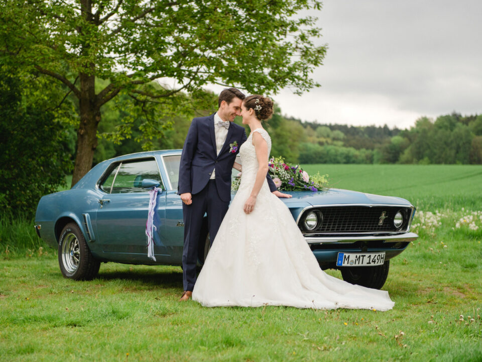 Ein Paar im Hochzeitskleid steht neben einem Oldtimer auf einer Wiese, im Hintergrund sind ein Baum und Grün zu sehen.