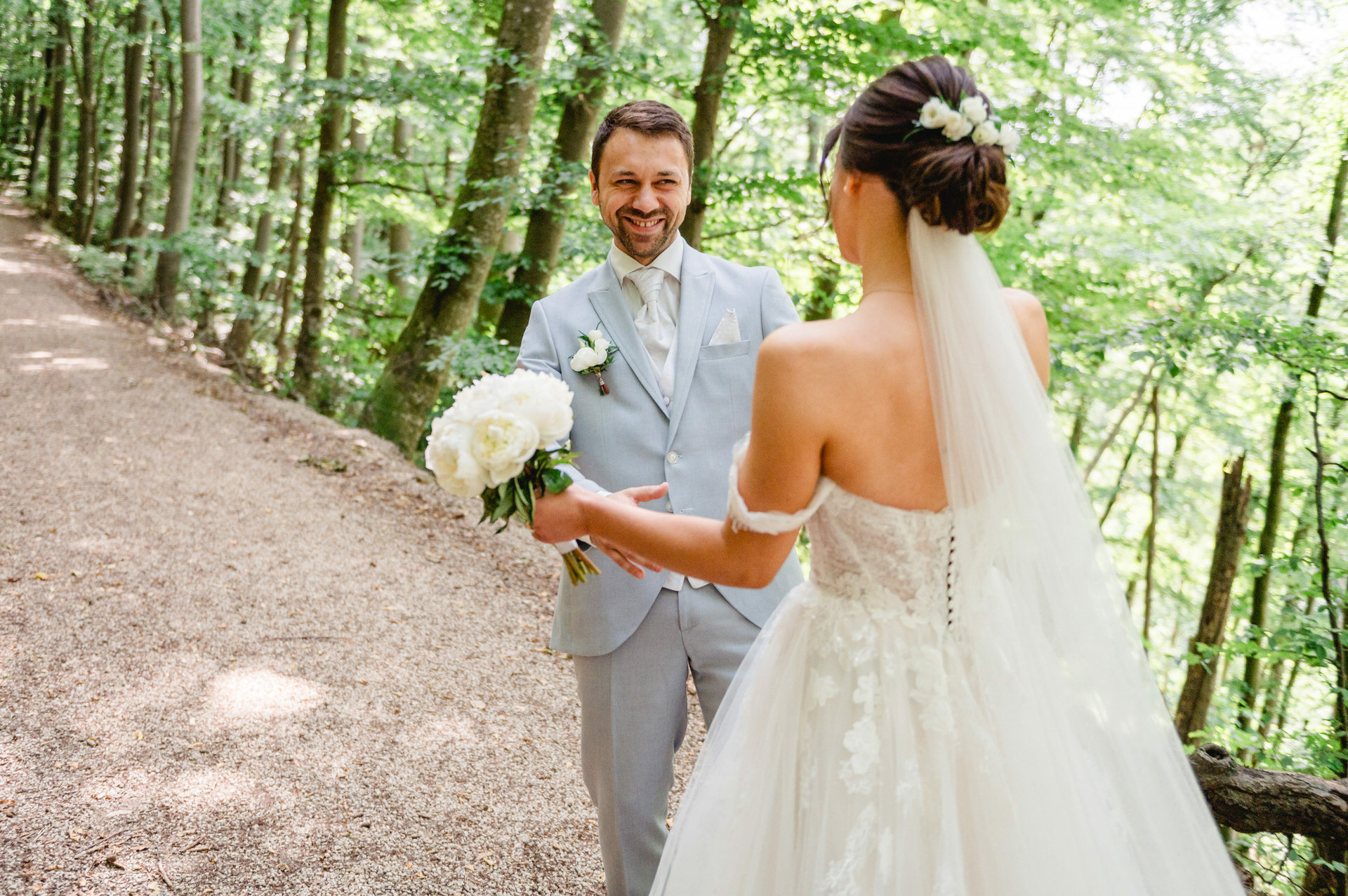 Eine Braut im weißen Kleid hält einen Blumenstrauß und steht auf einem Waldweg einem Bräutigam im hellgrauen Anzug gegenüber.