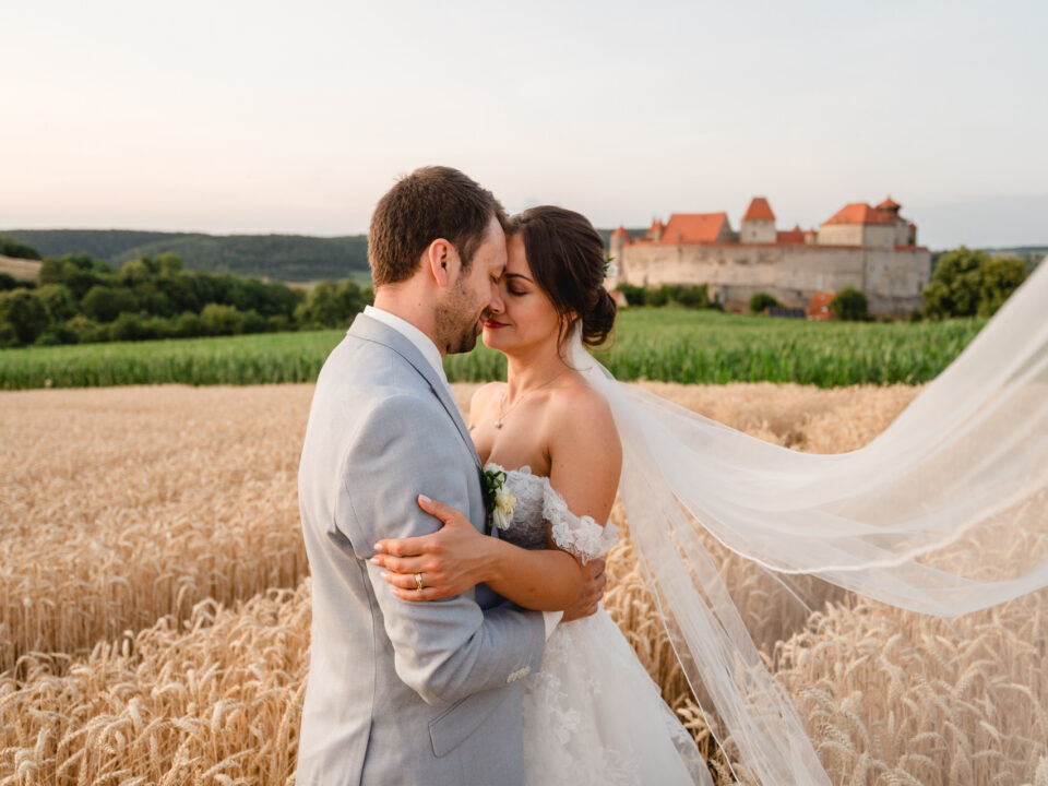 Braut und Bräutigam umarmen sich in einem Weizenfeld mit einem Schloss im Hintergrund. Der Schleier der Braut weht hinter ihr her.